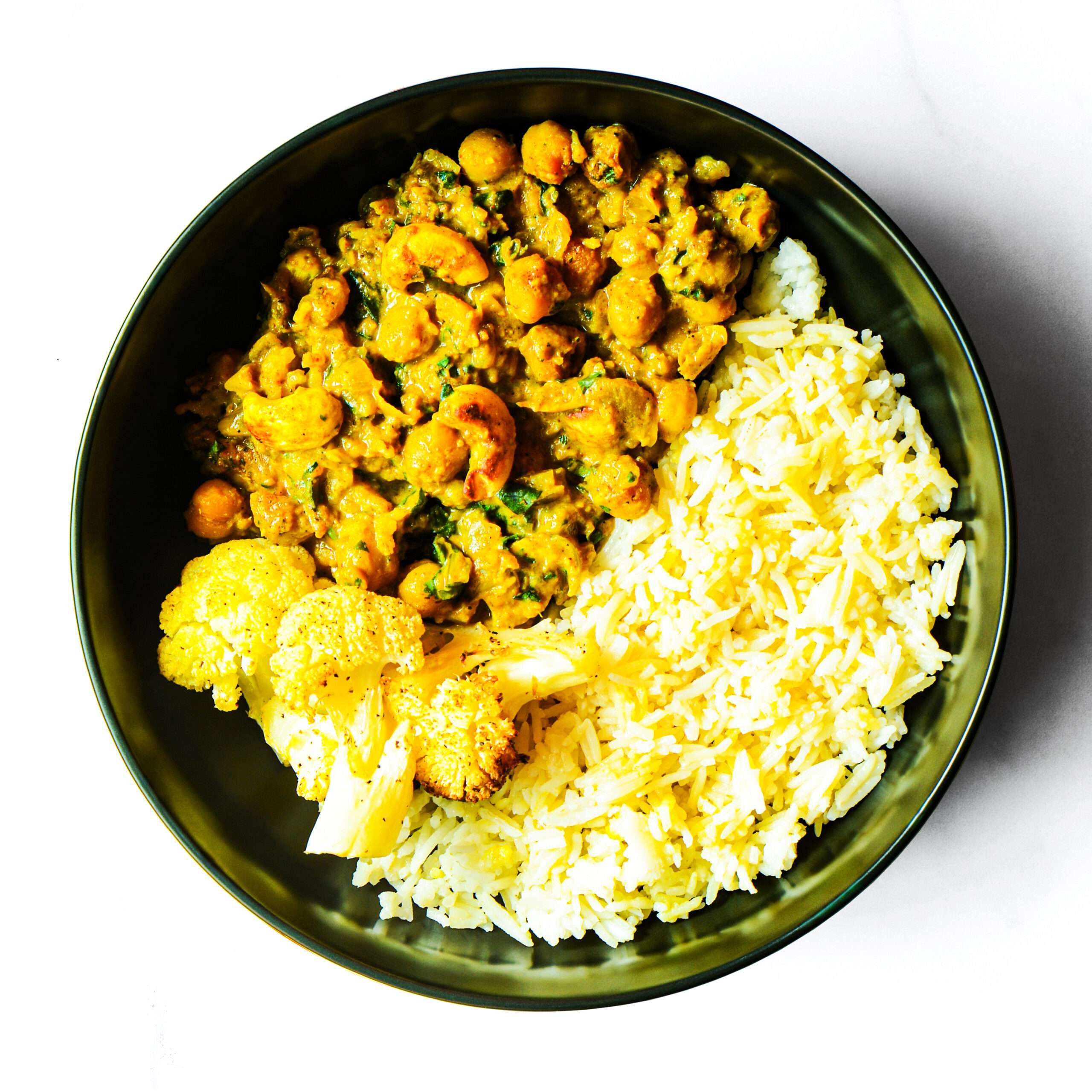 A black bowl containing yellow rice, roasted cauliflower, and a chickpea and vegetable curry, photographed from above on a white background.