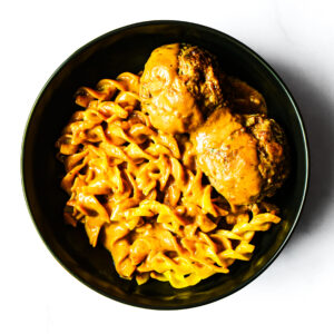 A black bowl filled with pasta in a creamy orange sauce, served with two large meatballs, photographed from above on a white background.