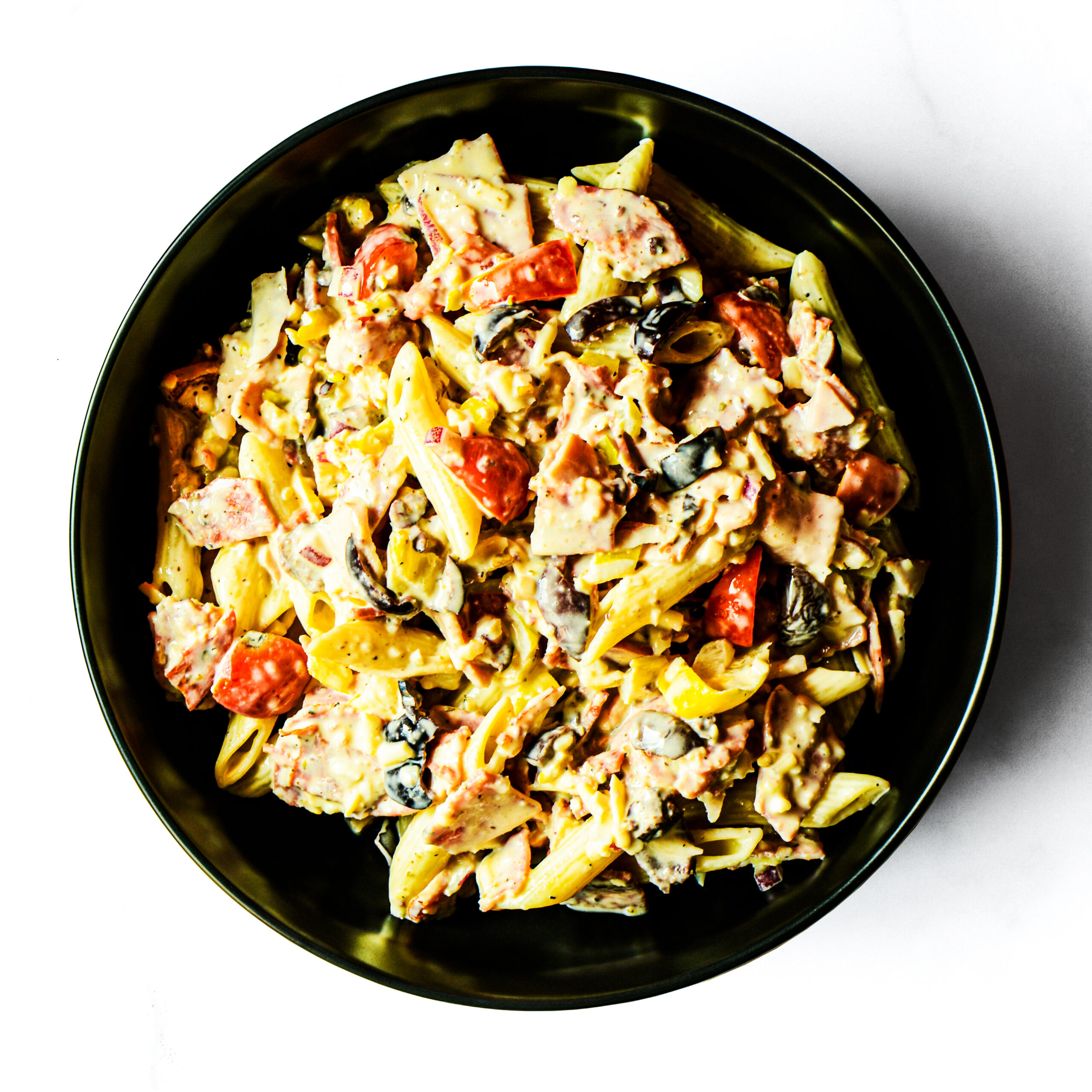 A black bowl filled with pasta mixed with sliced meat, tomatoes, olives, and a creamy sauce, viewed from above on a white background.